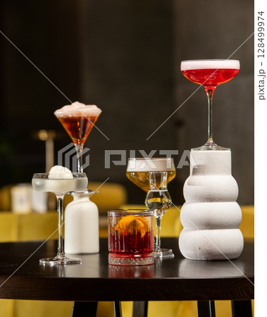 A richly colored red cocktail with a vibrant orange peel garnish is presented in a textured rocks glass, surrounded by other sophisticated drinks in a dimly lit bar setting. A richly colored red cocktail with a vibrant orange peel garnish is presented in a textured rocks glass, surrounded by other sophisticated drinks in a dimly lit bar setting. 128499974