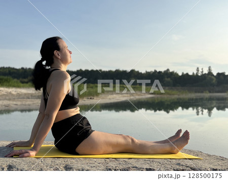 Adult woman sitting on yellow exercise mat and doing yoga outdoors by lake and forest. Summer fitness, sport activity in nature, healthy lifestyle, body care and mental balance Adult woman sitting on yellow exercise mat and doing yoga outdoors by lake and forest. Summer fitness, sport activity in nature, healthy lifestyle, body care and mental balance 128500175