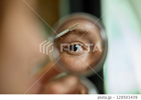 Close-up of woman eye applying brow gel seen through mirror with focused expression Close-up of woman eye applying brow gel seen through mirror with focused expression 128501439