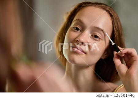Young woman smiling while applying clear brow gel with natural expression and ease Young woman smiling while applying clear brow gel with natural expression and ease 128501440