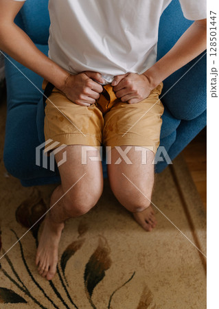 Vertical shot of unhealthy young man sitting on blue sofa grappling with urinary incontinence, anxiously clutching wet pants as navigates discomfort and embarrassment of situation, close-up. 128501447