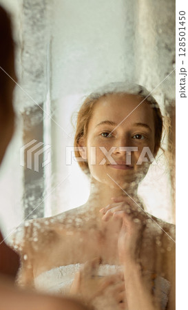 Skin-natural. Young woman gazing in foggy mirror with thoughtful emotion and soft light reflections 128501450