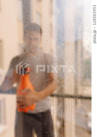 Vertical portrait of cleaning service employee washing window with spray bottle, viewed through glass adorned with water drops, creates soft blurred effect that enhances scene freshness and clarity. Vertical portrait of cleaning service employee washing window with spray bottle, viewed through glass adorned with water drops, creates soft blurred effect that enhances scene freshness and clarity. 128501451