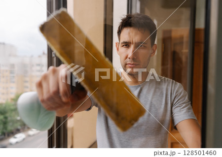 Focused young man washing window using squeegee and detergent, ensuring spotless and streak-free finish in residential home setting. Concept of domestic work, housekeeping and household. 128501460
