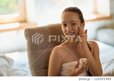 Young woman smiling while applying facial cream in relaxed morning skincare routine 128501488