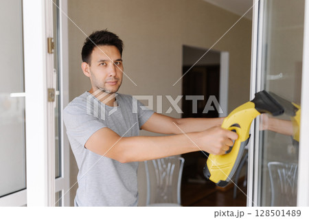 Portrait of handsome young man wiping glass surface using cordless handheld vacuum, working in bright contemporary living space, looking at camera. Concept of domestic work, housekeeping and household 128501489