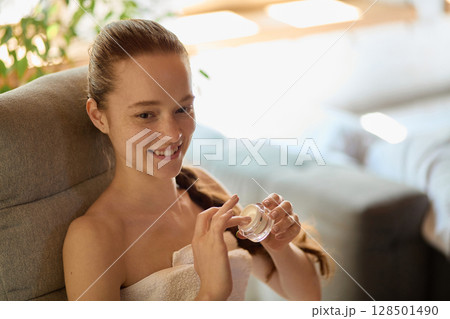 Smiling young woman applying skincare cream with joyful expression in cozy room Smiling young woman applying skincare cream with joyful expression in cozy room 128501490