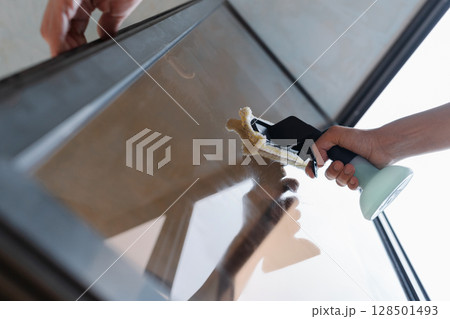 Close-up low-angle view of professional cleaning worker wiping large glass window, applying cleaning solution with spray bottle, using squeegee for spotless, streak-free result. Concept of household. Close-up low-angle view of professional cleaning worker wiping large glass window, applying cleaning solution with spray bottle, using squeegee for spotless, streak-free result. Concept of household. 128501493