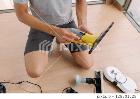 Close-up of unrecognizable service employee kneeling on floor, preparing cordless window cleaning devices and robot vacuum cleaner, organizing tools for efficient and thorough house cleaning routine. Close-up of unrecognizable service employee kneeling on floor, preparing cordless window cleaning devices and robot vacuum cleaner, organizing tools for efficient and thorough house cleaning routine. 128501529