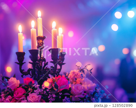 A close-up of a candelabra with lit white candles next to a bouquet of pink and white flowers, all set against a blurry purple background with bokeh lights. 128502890