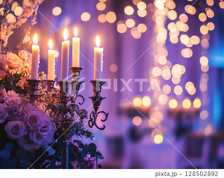 A close-up of a candelabra with lit white candles next to a bouquet of pink and white flowers, all set against a blurry purple background with bokeh lights. 128502892