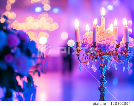 A close-up of a candelabra with lit white candles next to a bouquet of pink and white flowers, all set against a blurry purple background with bokeh lights. 128502894