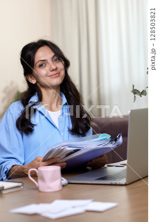 portrait of woman in blue shirt holds folders uses laptop in home office 128503821