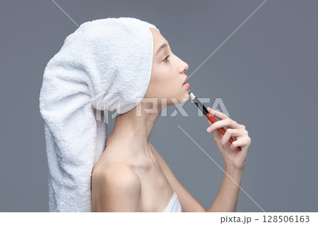 Young caucasian female applying makeup with brush after shower on grey 128506163