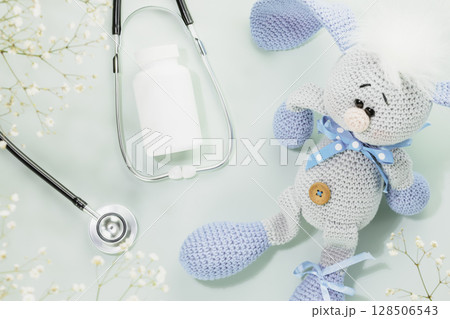 Kids health care. Kid supplements, vitamins or pils in white bottle with stethoscope and cute knitted toy rabbit on blue background with white flowers. Family doctor or pediatrician concept. Flat lay 128506543