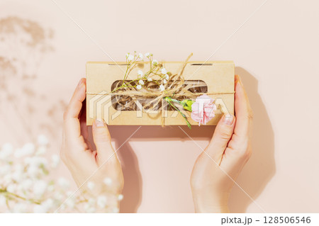 Female hands holding craft gift box with tender flowers on pink background. Romantic holiday card. Packaging and delivery concept. Flat lay, copy space 128506546