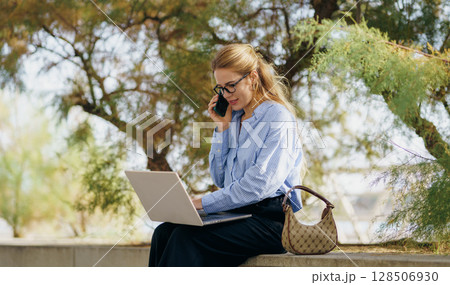 Professional Woman Successfully Working Outdoors with Her Laptop and Phone in Hand 128506930