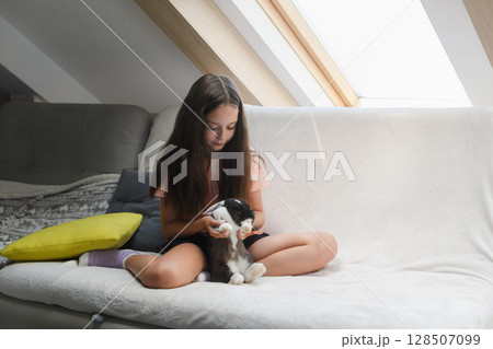 Smiling girl playing with black and white rabbit indoors. 128507099