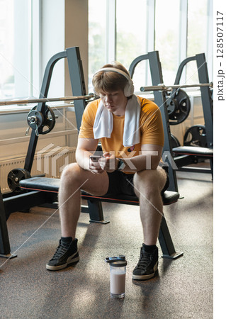 Young caucasian sporty man relax and sitting with mobile phone, protein shaker, listen music headphones at gym. Bodybuilder during training. Fitness lifestyle concept. Take a break. Vertical. 128507117