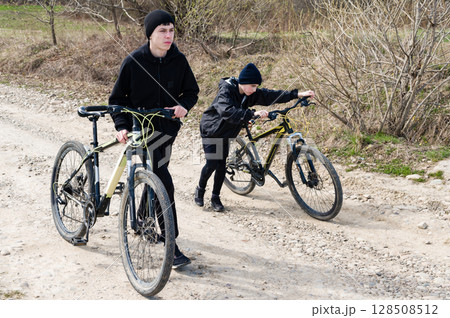 Two teens walk their bikes down a dirt road, one pushing his bike while the other walks alongside. 128508512