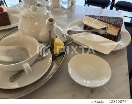 Piece of chocolate cake on white plate with tea bag in cup and tea-pot, lemon near it, sweet dessert in cafe, side view close up, selective focus 128508744