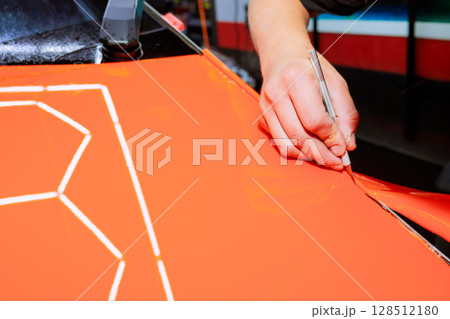 Skilled hand meticulously cuts through vibrant orange material using crafting knife inside car service 128512180