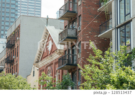 A red-brick church roof with classical trim stands beside old and new apartment buildings. Balconies, fire escapes, and high-rises contrast in this layered urban cityscape. 128513437