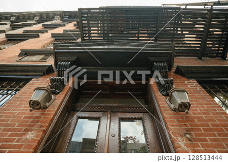 An upward perspective reveals a vintage brick building with ornate black trim, lantern lights, and a metal fire escape. The double wooden doors and classical detailing emphasize historic architecture. 128513444