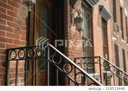 A close-up view of a decorative black iron stair railing leads up to a vintage wooden door. The red brick facade and wall lanterns enhance the building's classic New York charm. 128513446