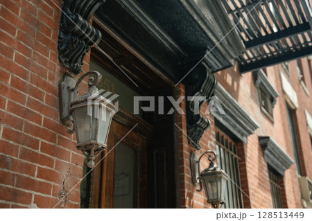 Two classic wall lanterns are mounted on a red brick facade, framed by black carved corbels and an ornate entrance. A metal fire escape casts shadows along the traditional architecture. Two classic wall lanterns are mounted on a red brick facade, framed by black carved corbels and an ornate entrance. A metal fire escape casts shadows along the traditional architecture. 128513449