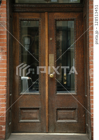 A detailed close-up of a vintage wooden double door with glass inserts, set in a red brick wall. The craftsmanship features carved trim and a brass handle with classic charm. A detailed close-up of a vintage wooden double door with glass inserts, set in a red brick wall. The craftsmanship features carved trim and a brass handle with classic charm. 128513451