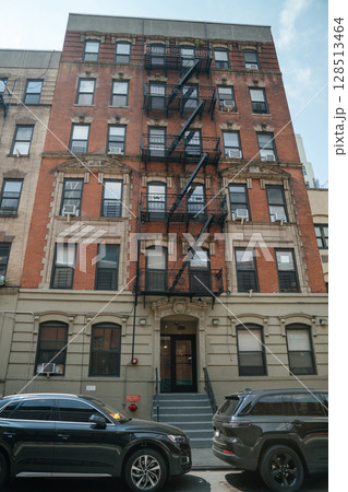 Multi-story brick apartment building features an exterior fire escape, set against a quiet city street. Two black cars parked in front enhance the somber mood of this urban scene during daytime. 128513464
