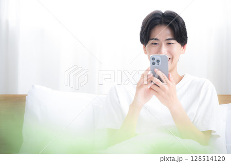 ベットでくつろぐスマホを持つ考える爽やかな男性の上半身　タップや画面を見る	 128514120