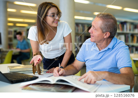 Happy casual Latin woman in glasses talking with older man reading in the library 128516076