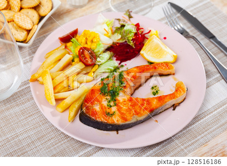 Salmon is seasoned with pesto and garnished with French fries, salad of fresh vegetables, crackers 128516186
