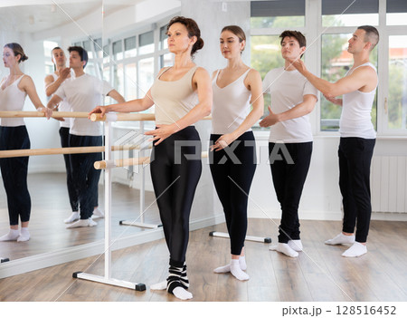 Group of young dancers engaged in ballet in dance studio perfoms exercise near ballet barre, male choreographer helps to coordinate movements correctly 128516452