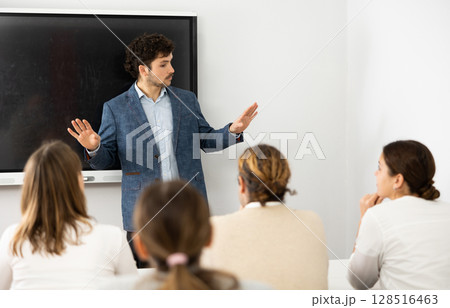 Male business coach standing near interactive board explaining topic while standing against group of people sitting at desks in front of him Male business coach standing near interactive board explaining topic while standing against group of people sitting at desks in front of him 128516463