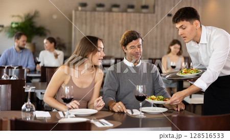 Male waiter serving fresh vegetable salads to married couple guests visiting restaurant for dinner, sitting at table with glasses of wine 128516533