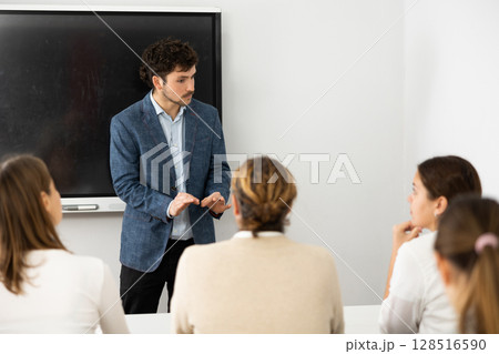 Male business coach standing near interactive board explaining topic while standing against group of people sitting at desks in front of him Male business coach standing near interactive board explaining topic while standing against group of people sitting at desks in front of him 128516590