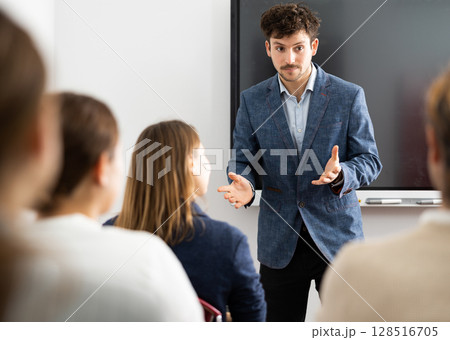 Young man standing near interactive board and communicating with adult students during advanced training courses in classroom Young man standing near interactive board and communicating with adult students during advanced training courses in classroom 128516705