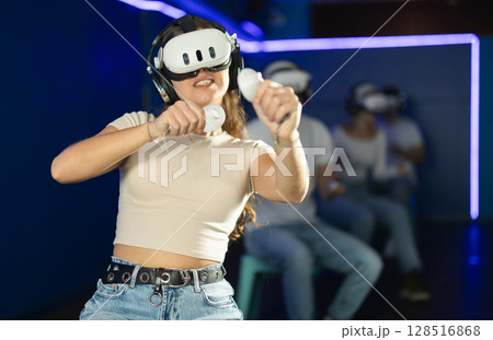 In darkened hall of gaming club, girl in VR helmet takes part in group game In darkened hall of gaming club, girl in VR helmet takes part in group game 128516868
