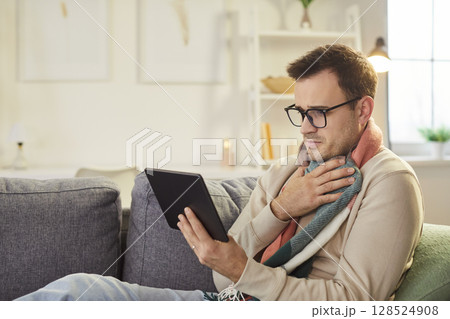 Sick young man sitting on sofa at home using tablet with seasonal flu. Sick young man sitting on sofa at home using tablet with seasonal flu. 128524908
