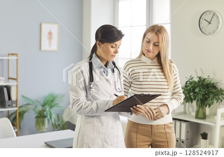 Female doctor in lab coat with clipboard explaining medical details while consulting young woman. 128524917
