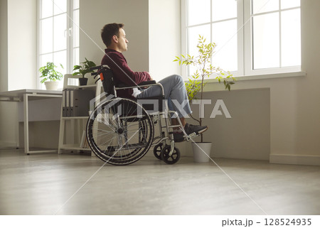 Disabled man sitting in wheelchair and looking through window, being on rehabilitation 128524935