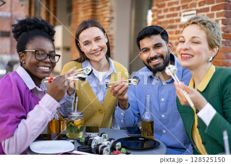 A diverse group of friends enjoys sushi together, smiling and laughing while eating outdoors on a sunny day. 128525150