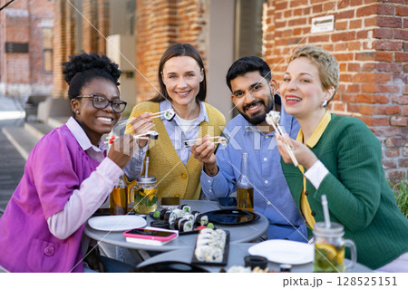 A diverse group of friends enjoys a sushi meal outdoors, smiling and using chopsticks at a table. 128525151