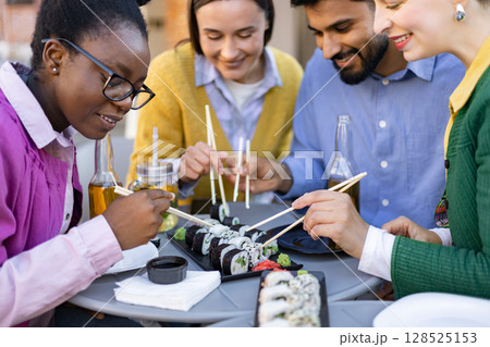 A diverse group of friends enjoys a sushi meal together at an outdoor restaurant, using chopsticks. 128525153