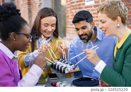 Diverse group of friends enjoys sushi and drinks together at an outdoor restaurant. 128525220