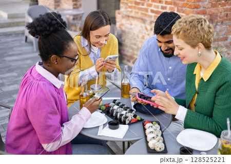 A diverse group of friends enjoy sushi and drinks while using their smartphones at an outdoor cafe. 128525221