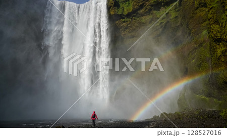 Unidentified woman stands beneath the powerful Skogafoss waterfall in Iceland, framed by mist and a vivid rainbow 128527016